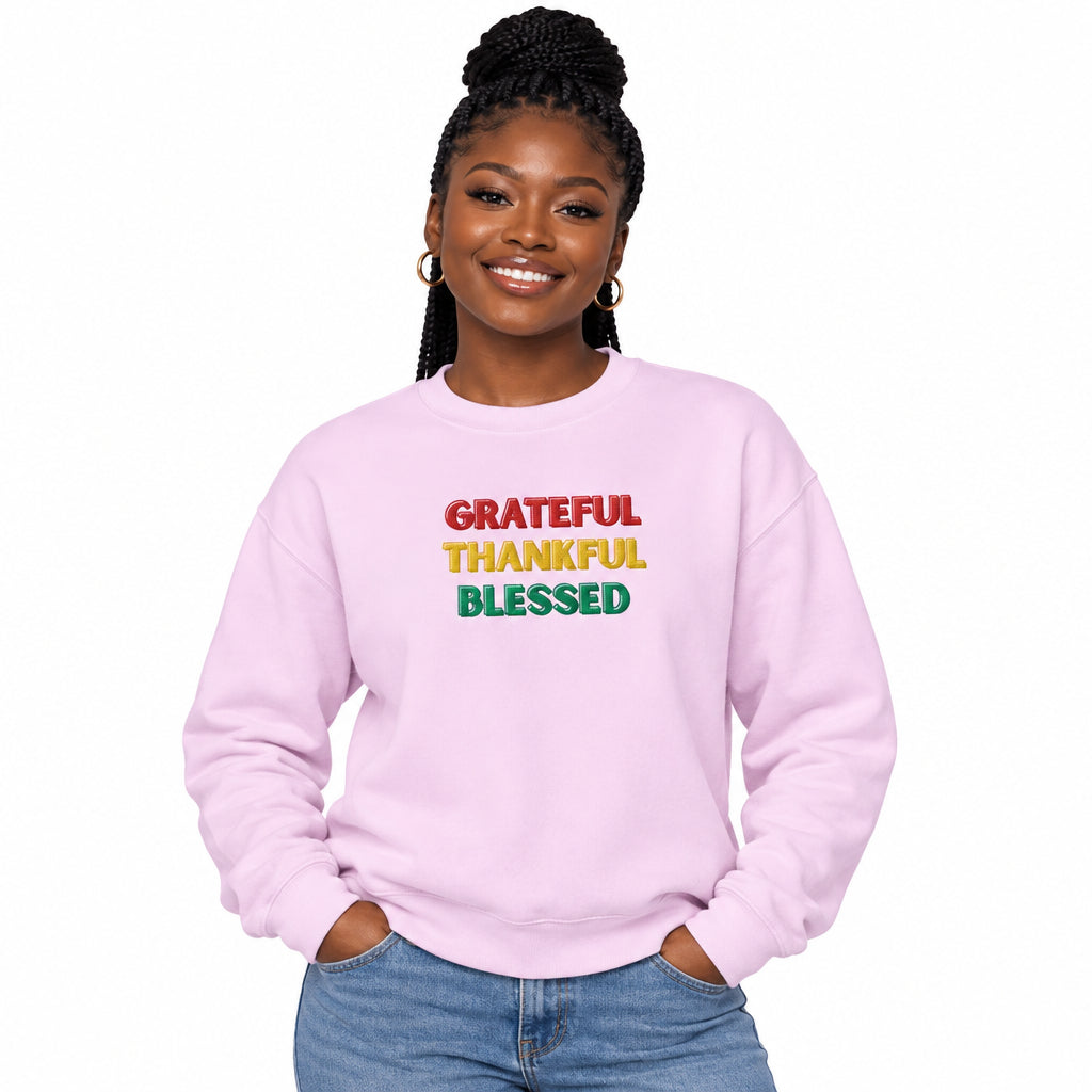 A light pink crewneck sweatshirt with the words 'GRATEFUL', 'THANKFUL', and 'BLESSED' printed in red, yellow, and green block letters on the chest, shown against a plain white background.