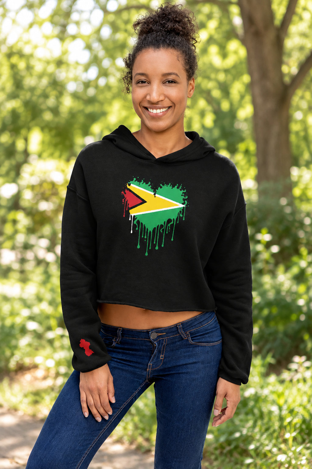 A woman wearing a black cropped hoodie with a circular graphic of a profile portrait and a Guyana flag motif on the chest, paired with blue jeans, against a plain white background.