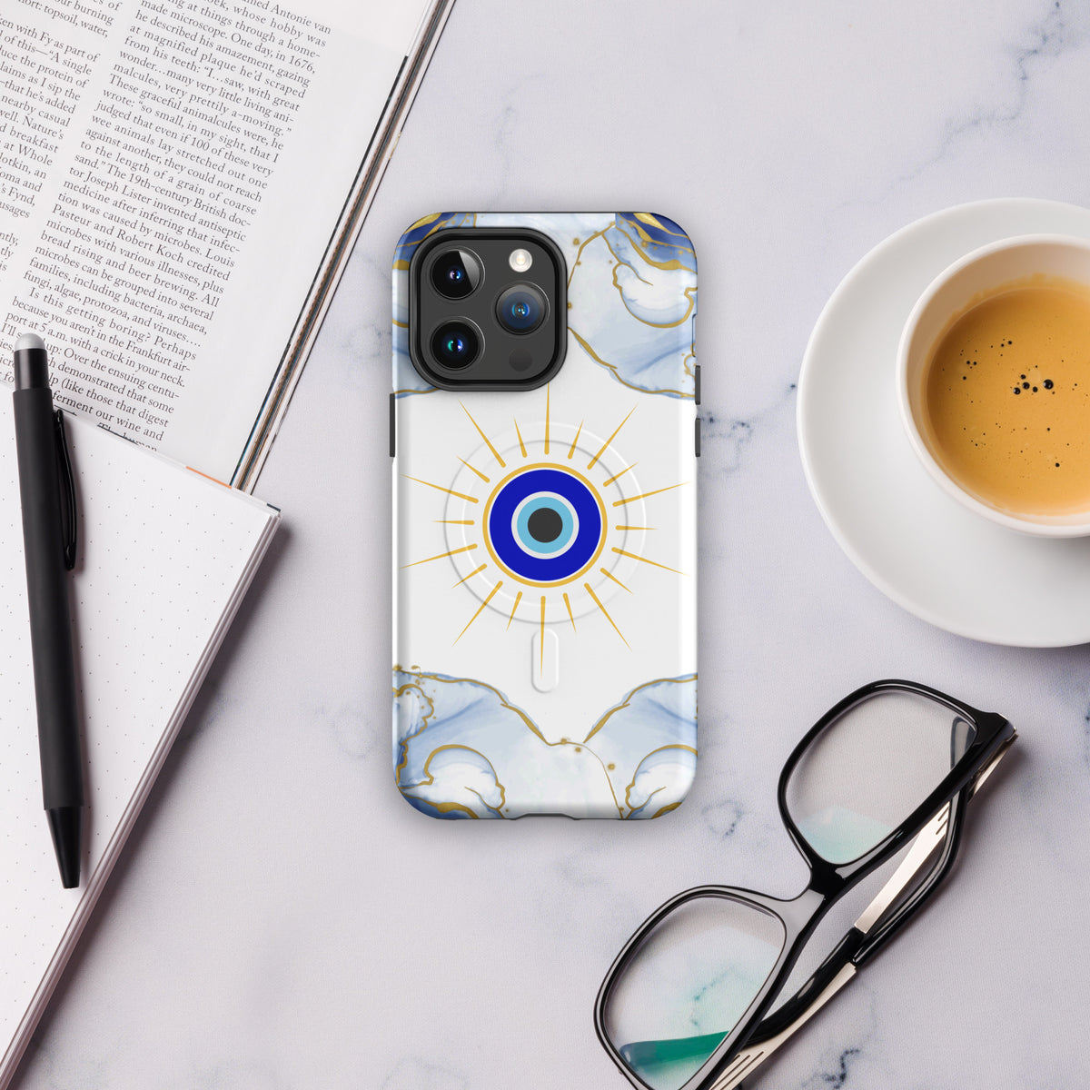 Phone case with sun evil eye design on a marble surface with a book, pen, and coffee cup.