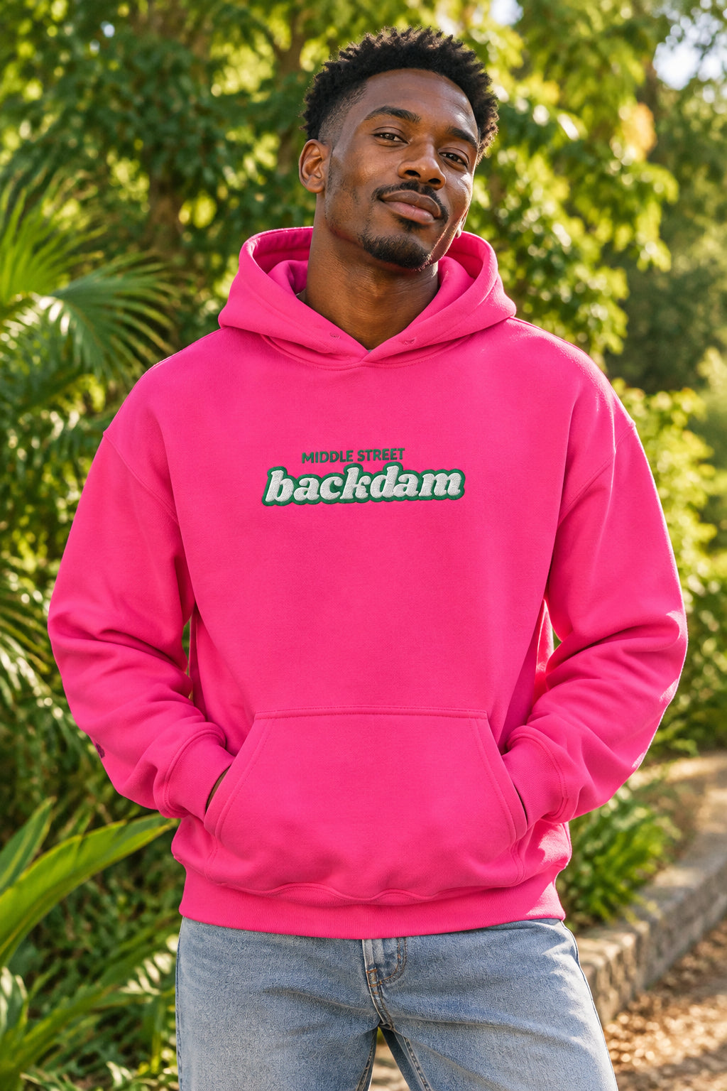 A bright pink pullover hoodie with a front kangaroo pocket and a chest graphic reading 'MIDDLE STREET backdam' in teal and white. A small black graphic is visible on the left sleeve.
