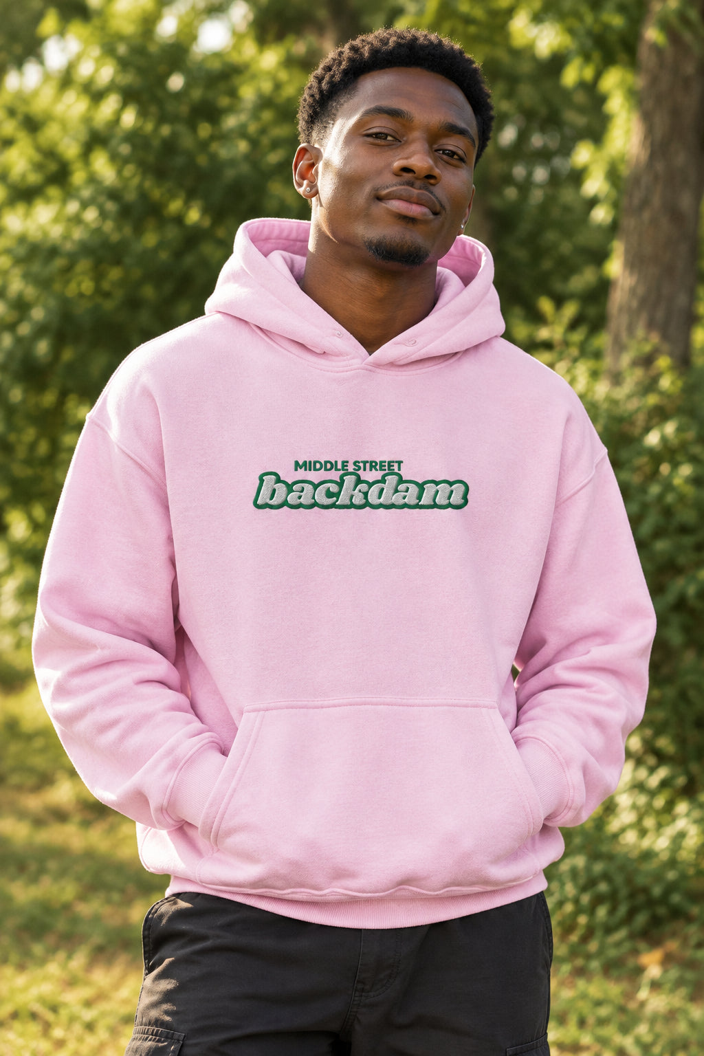 A light pink pullover hoodie with a front kangaroo pocket and a chest graphic reading 'MIDDLE STREET backdam' in green and white. A small black Guyana map graphic is visible on the left sleeve cuff.