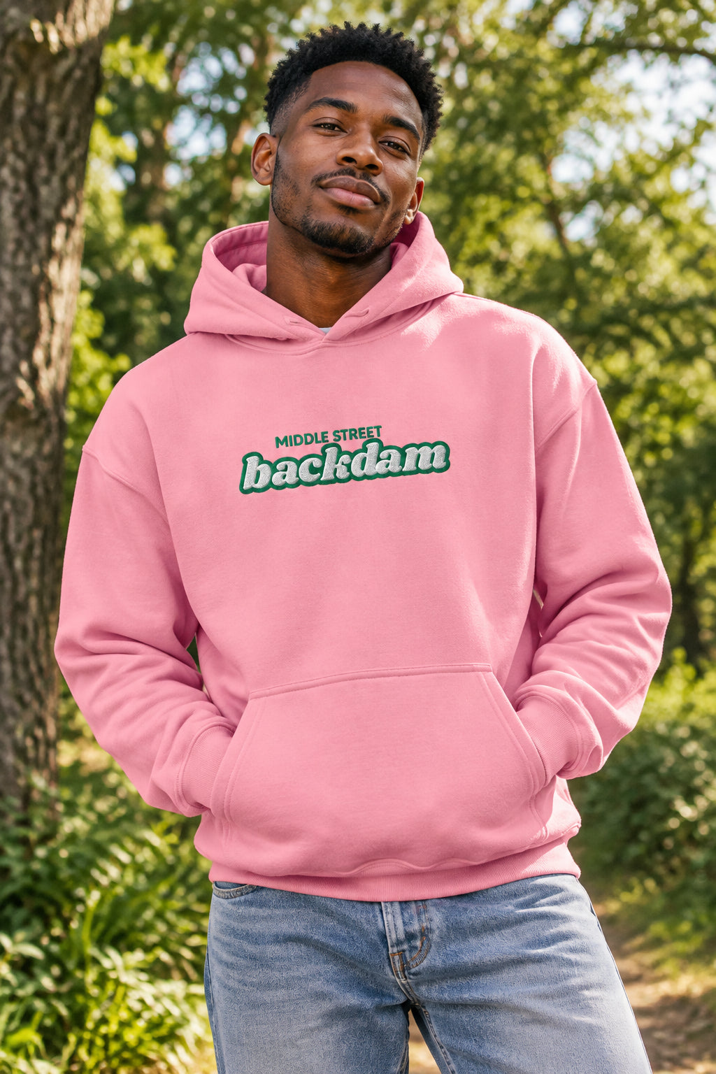 A pink pullover hoodie with a front kangaroo pocket and a chest graphic that reads 'MIDDLE STREET backdam' in green and white. A small black graphic is visible on the left sleeve cuff.